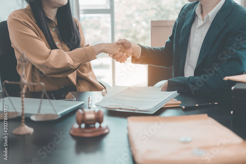 The lawyer and his client are shaking hands and discussing documents and ideas at a conference table. Both are wearing suits. Legal documents and a statue are placed on the table.