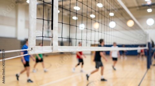 Volleyball net close up in focus at indoor gymnasium with blurred athletes practicing team sports in background during competitive match training session on wooden court floor