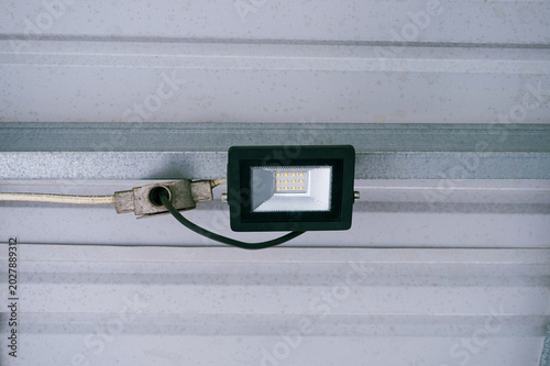 Industrial spotlight hangs from metal beams. White corrugated ceiling reflects bright, cool light. Metal framework supports modern lighting. Ideal for warehouses, factories, or industrial spaces