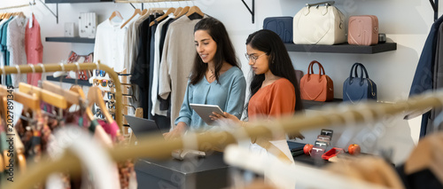 Two young women collaborate at checkout counter inside stylish clothing boutique, working on laptop using digital tablet. Teamwork, customer services, small business management in modern fashion store