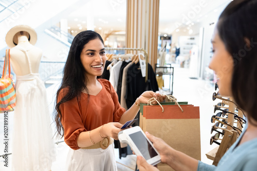 Happy young Indian woman customer holding two paper bags with goods, swiping her credit card to contactless payment terminal held by store clerk. Fast and secure cashless transactions in retail space