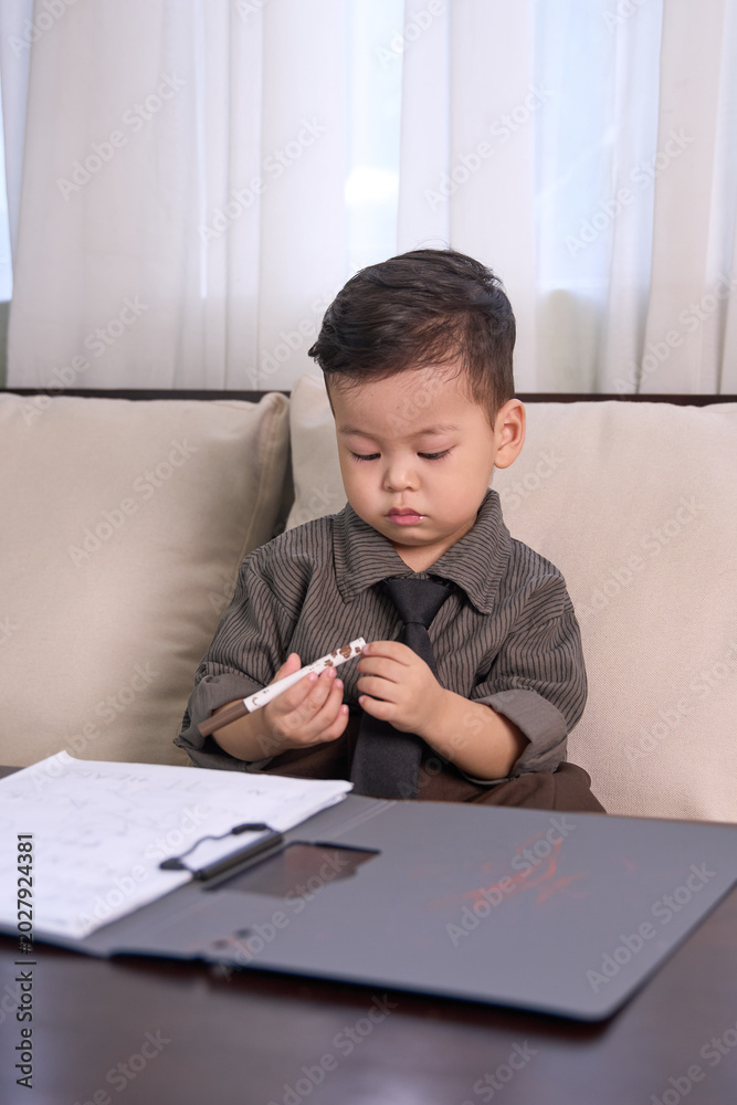 Naklejka premium A 2-year-old toddler boy sitting at the table holding a pen over the clipboard
