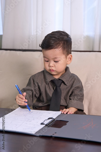 A 2-year-old little boy sitting on the sofa writing carefully with a blue pen
