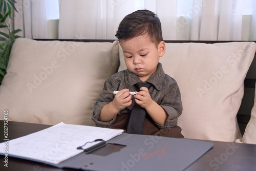 A 2-year-old baby boy sitting on the sofa looking down and playing with the pen