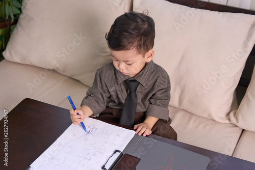 A 2-year-old toddler boy sitting at the table drawing with a pen on paper