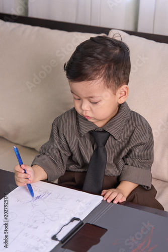 A 2-year-old baby boy sitting at the table writing with a blue pen on the clipboard