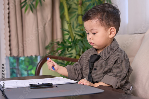 A 2-year-old toddler boy sitting on the sofa pointing with a pen at the paper