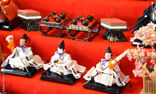 Traditional Japanese hina dolls arranged on a red tiered stand, featuring three male figures and miniature offerings of food and utensils