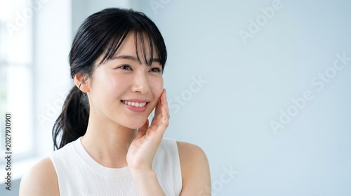 Young smiling Asian woman touching her glowing healthy skin in bright room.