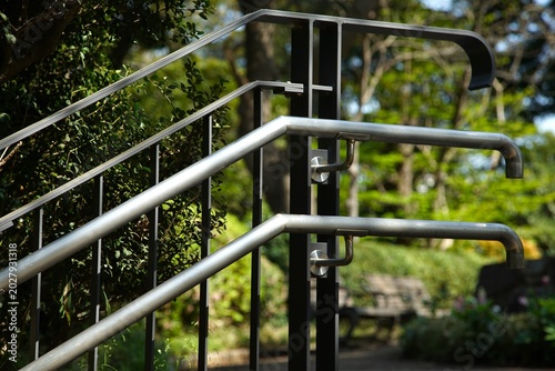 Metal handrails of different heights installed along a paved garden pathway, with green trees and plants visible in the background
