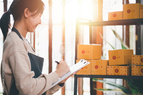 Entrepreneur checking inventory and managing online product orders in small warehouse workspace, supporting e-commerce fulfillment, stock organization, parcel preparation, retail shipping operations.