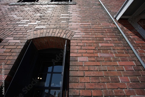 Sunlit red brick wall of an old industrial building, seen from below with an open black framed window