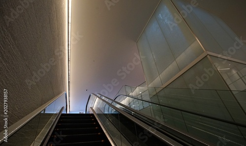 An upward view of an escalator in a modern interior, featuring glass side panels and a lit, frosted glass structure alongside a textured wall