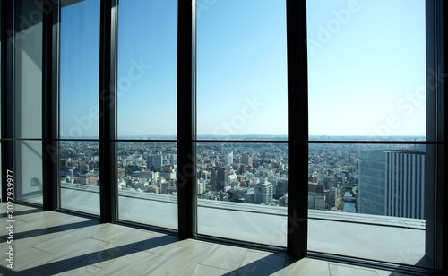 A wide-angle shot taken from the interior of a high-rise building, showing an expansive cityscape under a clear blue sky through large glass windows