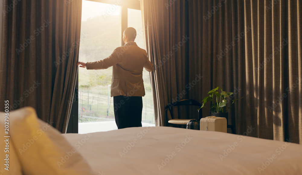 Fototapeta premium Man, sunlight and open curtain in hotel room for window view to start day, fresh air and relax. Business trip, accommodation and person at lodge with luggage, back and bedroom for hospitality