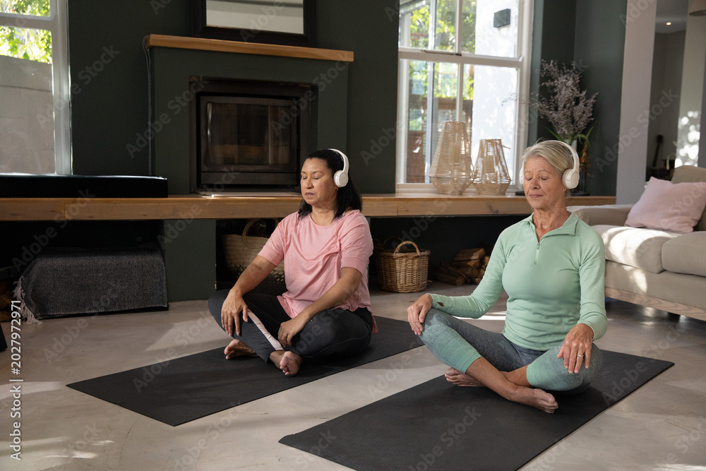 Fototapeta premium Mature, senior women friends meditating on black yoga mats at home in activewear wearing headphones
