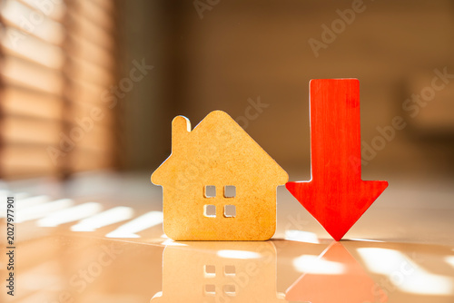 Real estate housing market decline concept. Wooden house model standing next to falling red arrow on floor with window blind shadows. Decreasing property value and mortgage rate.