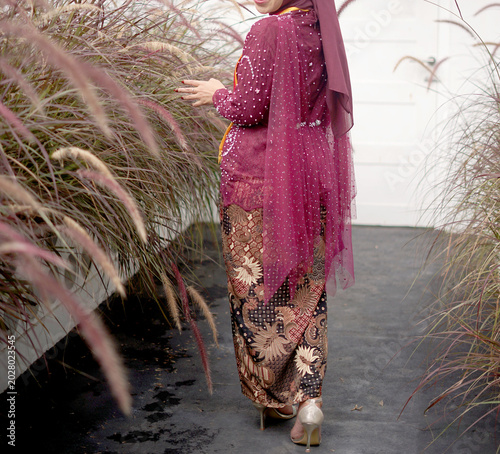 A faceless female body wearing a luxurious burgundy kebaya with beautiful batik cloth, standing in a garden of reeds with a white wall in the background