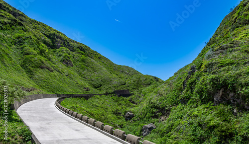 Winding white concrete road disappearing into green mountains under clear blue sky.