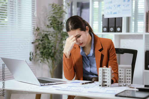 Stressed female real estate agent working on condominium and property sales project in office, feeling pressure while analyzing housing market documents.