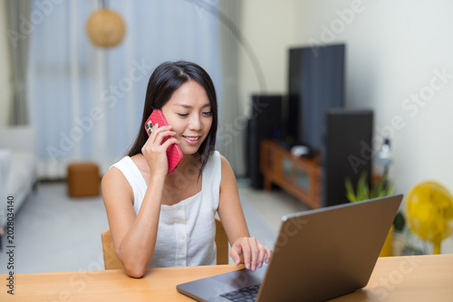 Smiling asian woman talking on mobile phone while using laptop computer