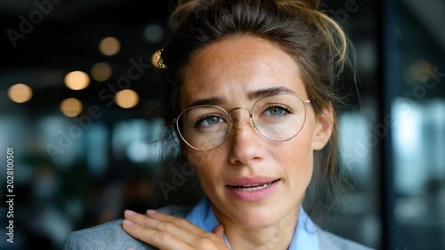 Concerned Woman in Office: A close-up portrait of a thoughtful woman wearing glasses, with her hand slightly raised, conveying a sense of worry or deep thought in a blurred office environment.