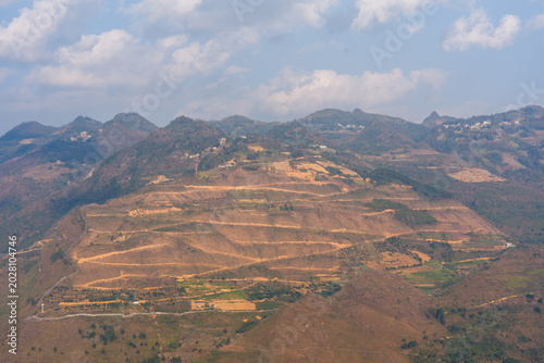 Rolling hills stretch under a bright sky. Terraced fields dot the rugged terrain. Sparse greenery contrasts with dry earth. Scattered trees dot the hillsides. Mountains rise in the distance, hazy