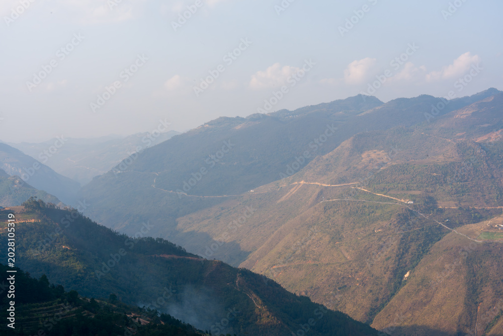 Obraz premium Mountain range stretches across the horizon. Winds stir the dry, sun-baked slopes. Distant ridges fade beneath a hazy sky. Faint trail winds along the valley's edge