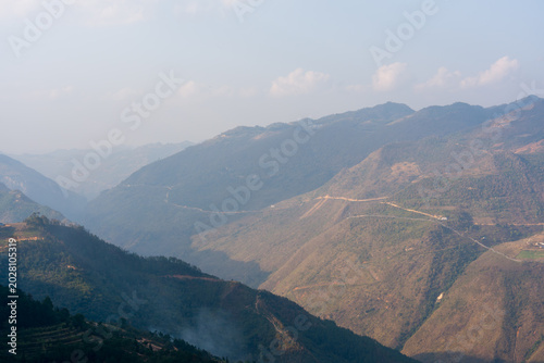 Mountain range stretches across the horizon. Winds stir the dry, sun-baked slopes. Distant ridges fade beneath a hazy sky. Faint trail winds along the valley's edge