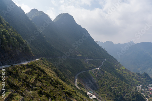 Winding road cuts through lush mountain slopes. Green hills stretch beneath a cloudy sky. Rugged peaks rise in the distance. Trail curves through dense forest terrain