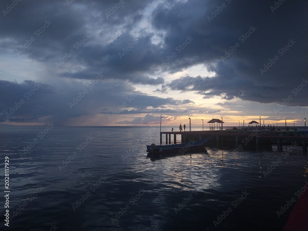 Obraz premium Dramatic Sunset Over the Ocean Pier with Boat and Stormy Clouds