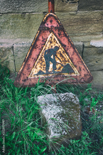 Old Rusty Road Construction Warning Sign Leaning Against Stone Wall, Abandoned and Overgrown
