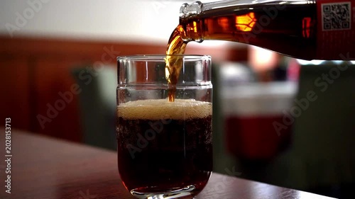 A close-up shot of a dark carbonated drink being poured from a bottle into a glass, creating a foamy head, set in a blurred warm restaurant atmosphere.