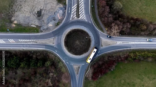 Aerial drone view from directly above of a public transport bus driving through a circular road intersection (roundabout) in a rural or suburban landscape.