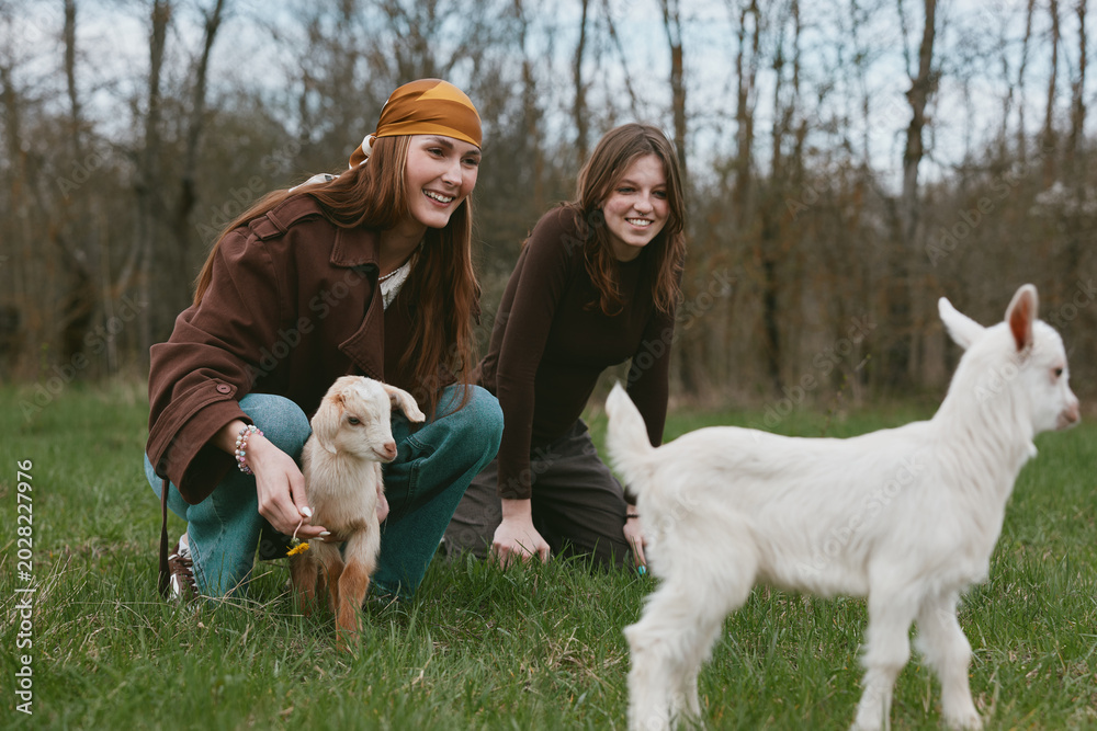 Fototapeta premium Two girls share a playful moment with a kid goat outdoors in the countryside, capturing a lifestyle scene with friends, animals and nature on a grassy field, bright smiles and candid interaction