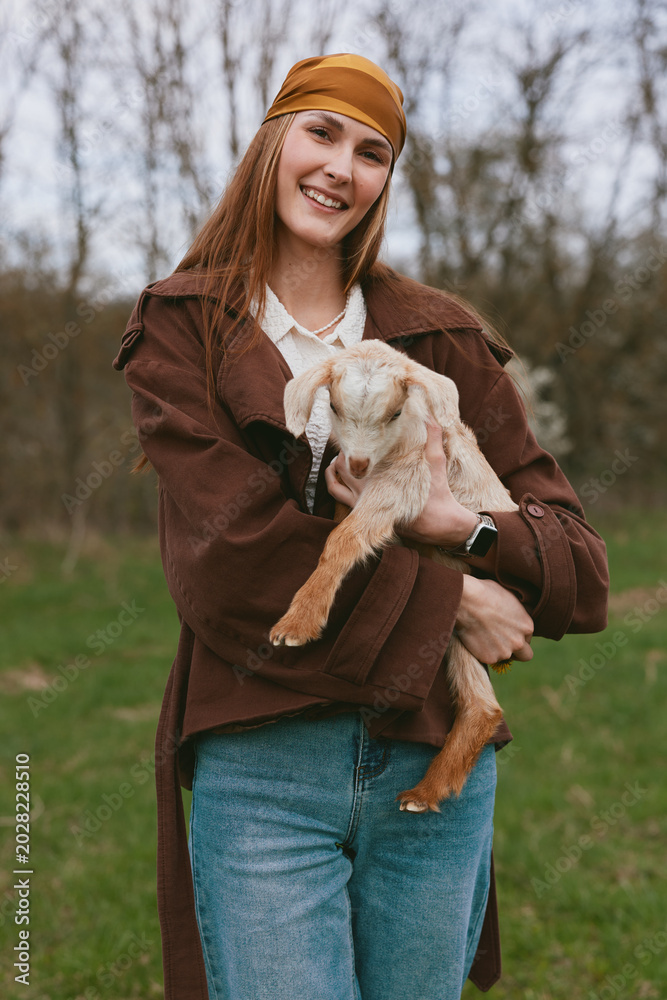 Fototapeta premium Joyful woman outdoors with a goat kid, sharing a warm moment in a rustic lifestyle setting. A candid scene of care, companionship, and natural farming vibes in a grassy field.