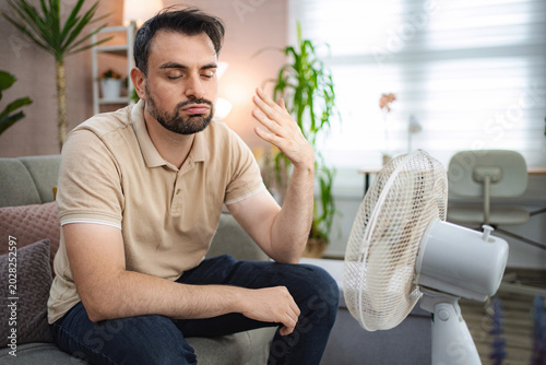 Fotografía Sweaty man cooling off with electric fan at home during summer heatwave