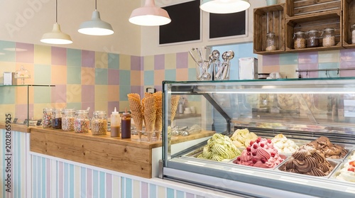 A vibrant ice cream parlor with a colorful tiled wall and a display case filled with various flavors of gelato and sorbet, including fruit and chocolate options, alongside waffle cones and toppings.