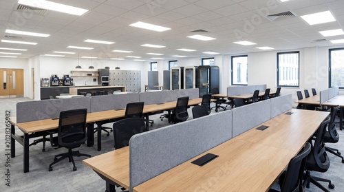 A modern, empty office space with rows of desks and chairs, illuminated by overhead lights. The interior features a minimalist design with grey dividers and wooden desktops.