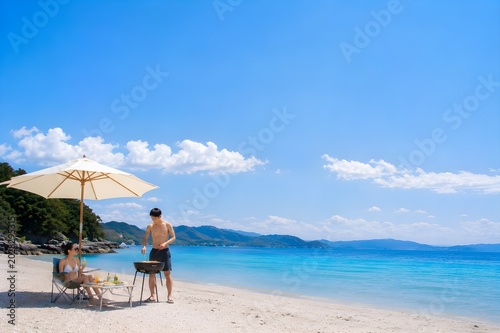 晴れた日のビーチでバーベキューを楽しむ男女の夏休みとアウトドアレジャーイメージ（AI生成）