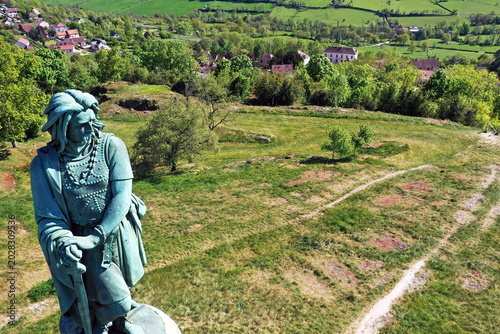 Εκτύπωση καμβά Das monumentale Vercingétorix-Denkmal auf einem steinernen Sockel, eingebettet in eine grüne Hügellandschaft mit Bäumen unter einem strahlend blauen Himmel