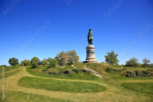 Quadro su tela Das monumentale Vercingétorix-Denkmal auf einem steinernen Sockel, eingebettet in eine grüne Hügellandschaft mit Bäumen unter einem strahlend blauen Himmel