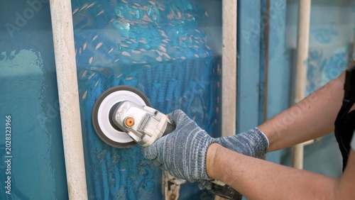Man in gloves uses an angle grinder with a wire brush attachment to remove old paint and rust from a blue metal shipping container. Close up on DIY restoration and surface preparation work