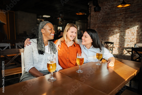 Fotografie Three diverse women friends enjoying beer and laughing in a pub