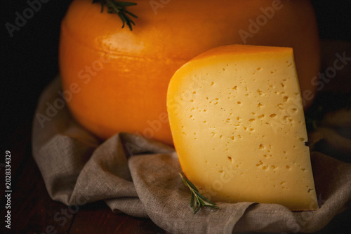 A close-up of wheels of cheese wrapped in yellow latex against a dark background. A piece of cheese lies nearby. A cross-section of the cheese. Cheese with holes. A farm product made from natural ingr