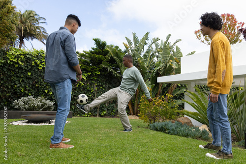 Diverse male friends playing soccer on backyard lawn, kicking ball near fire pit wearing sneakers