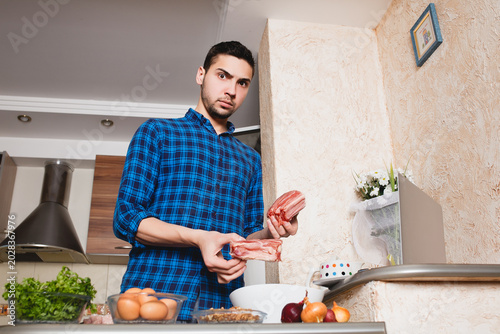 Fotografie Shocked or surprised young man holding raw pork ribs over a white bowl while preparing a meal in the kitchen