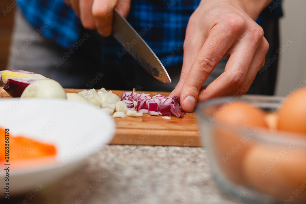 Obraz premium Man slicing red and white onions with a kitchen knife on a wooden board, culinary process concept with blurred ingredients.