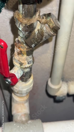 Corroded hot water meter connection leaking near red valve handle plumber inspecting closeup of calcified pipe joint with rust