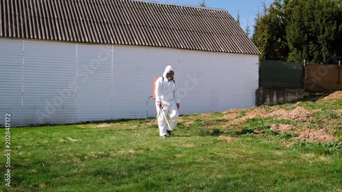 Person spraying pesticide on lawn field wearing a disposable protective suit. Treatment of grass from weeds and insects. Pest control. Insecticide sprayer with a proper protection. Garden care season.